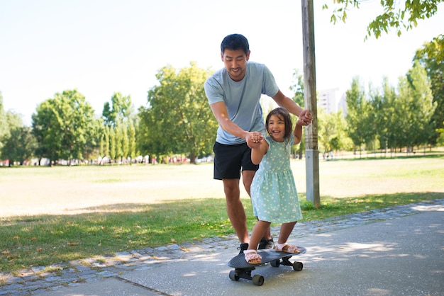 Easy family fitness activities l Portrait of Asian dad and little girl training to skateboard. Smiling man walking on alley holding daughters hands while she riding on skateboard. Active rest, healthy lifestyle and fatherhood concept